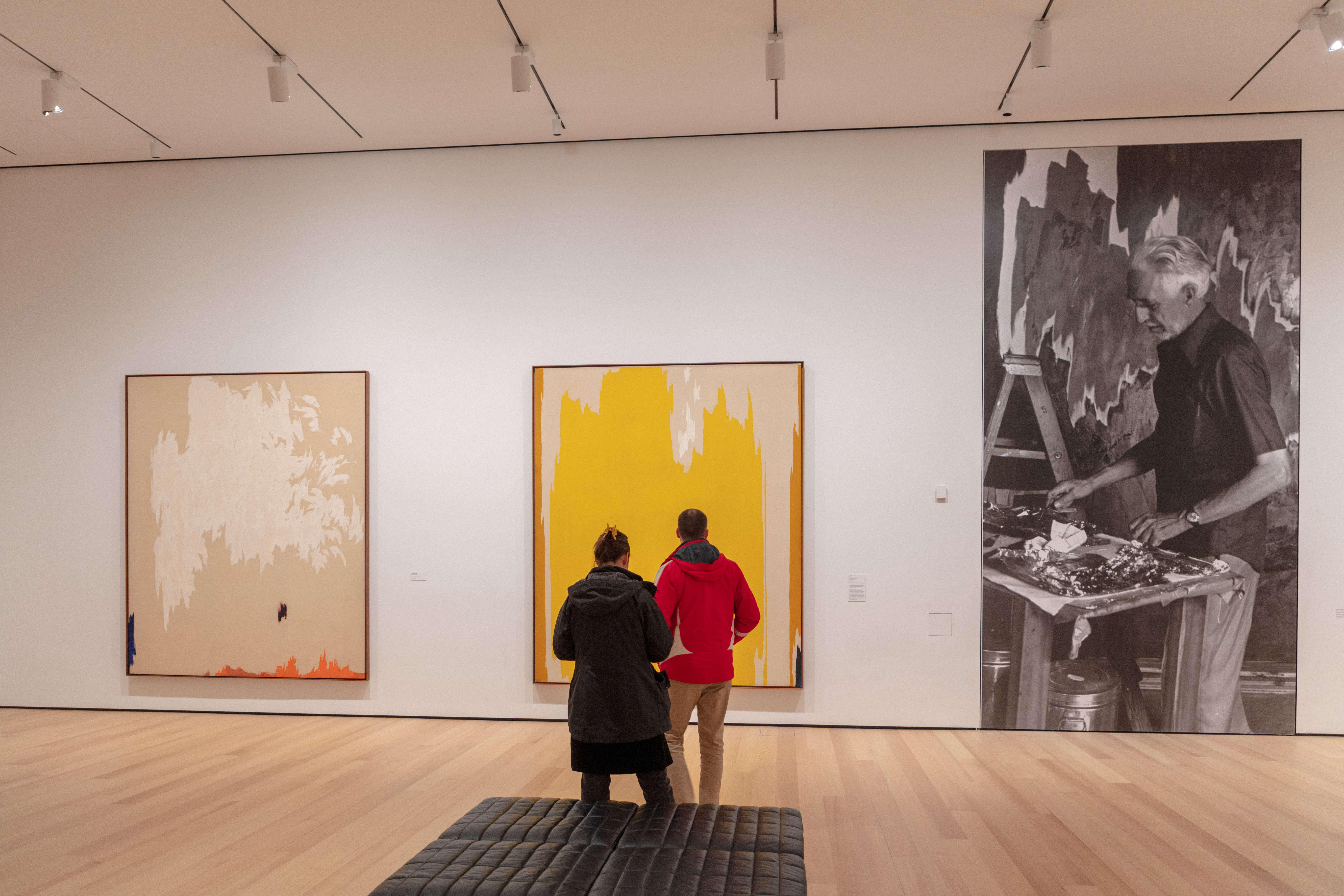 A man and woman in winter coats in the middle of an art gallery facing two large abstract paintings and large blow up photo of a man painting in a studio