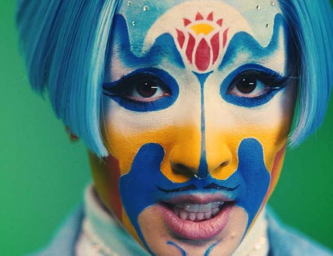 A person with a blue wig and facepaint with a red lotus, blue lines and yellow and red shading on the sides of their face 