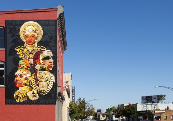 Photo of mural featuring three Black women adorned in futuristic gold crowns and jewelry against a black background on a red brick building, in the background bright blue sky