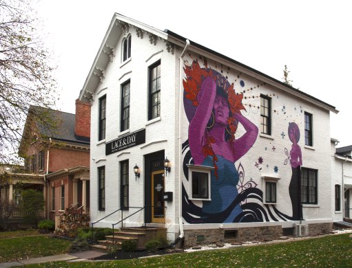 view of a white brick house on a residential street, with a mural of a purple-skinned woman in a blue dress holding her hands to her hair above her head