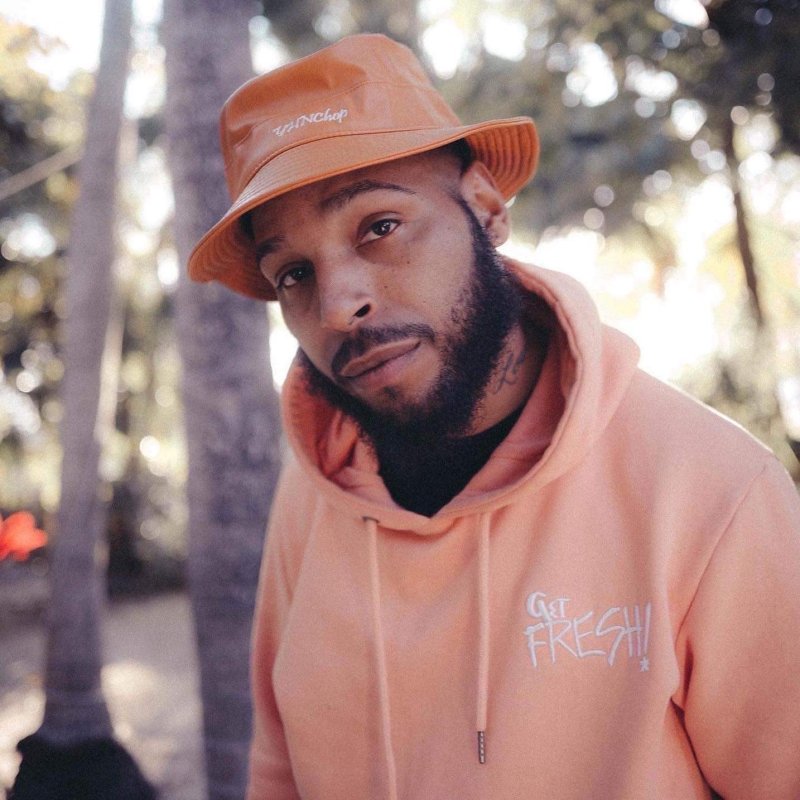 A man with dark facial hair and medium to dark skine tone wearing an orange hoodie and hat