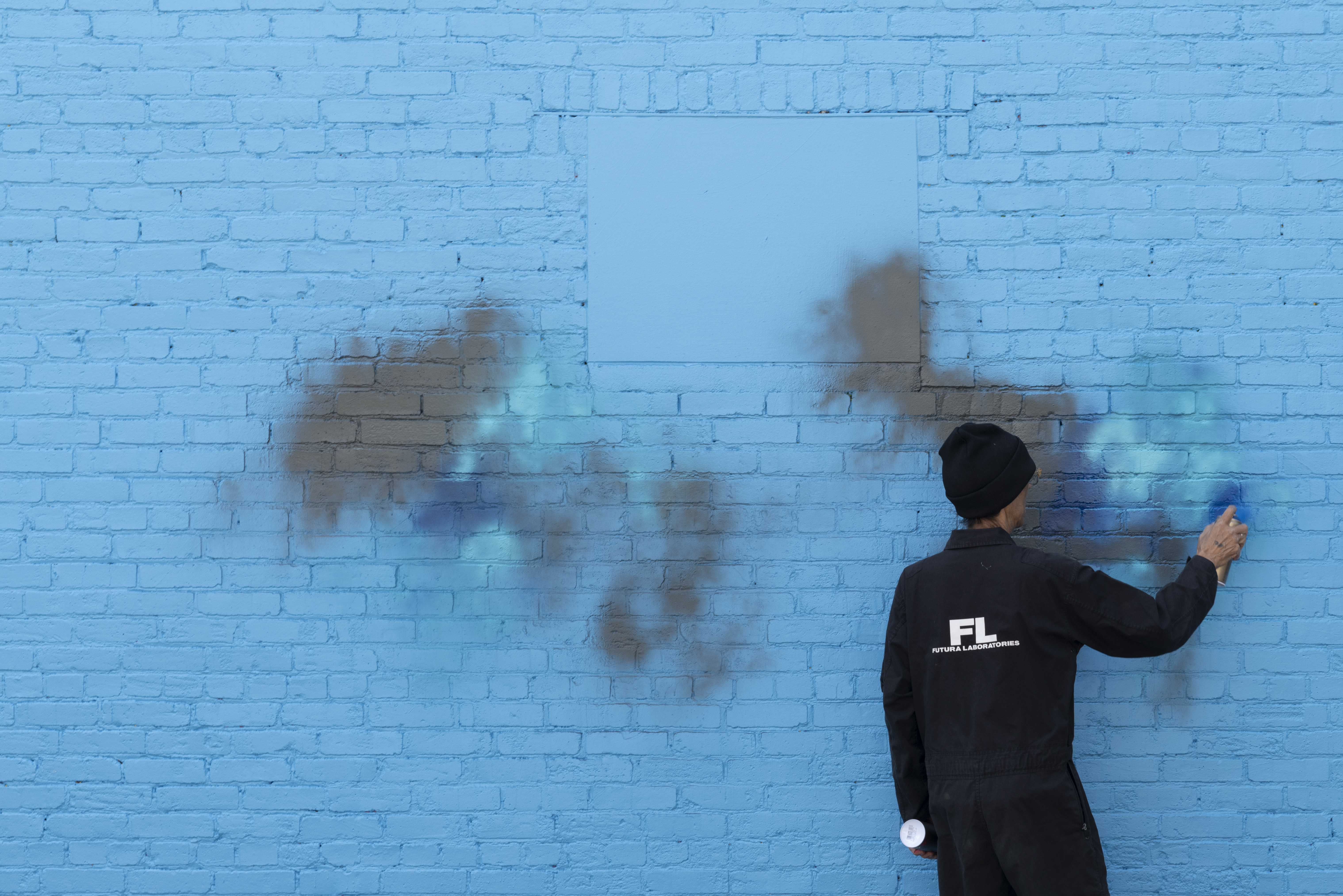 A man in a black beanie and black jumpsuit with the words FuturaLaboratories on the back holds spray paint can in his right hand up to a bright blue wall