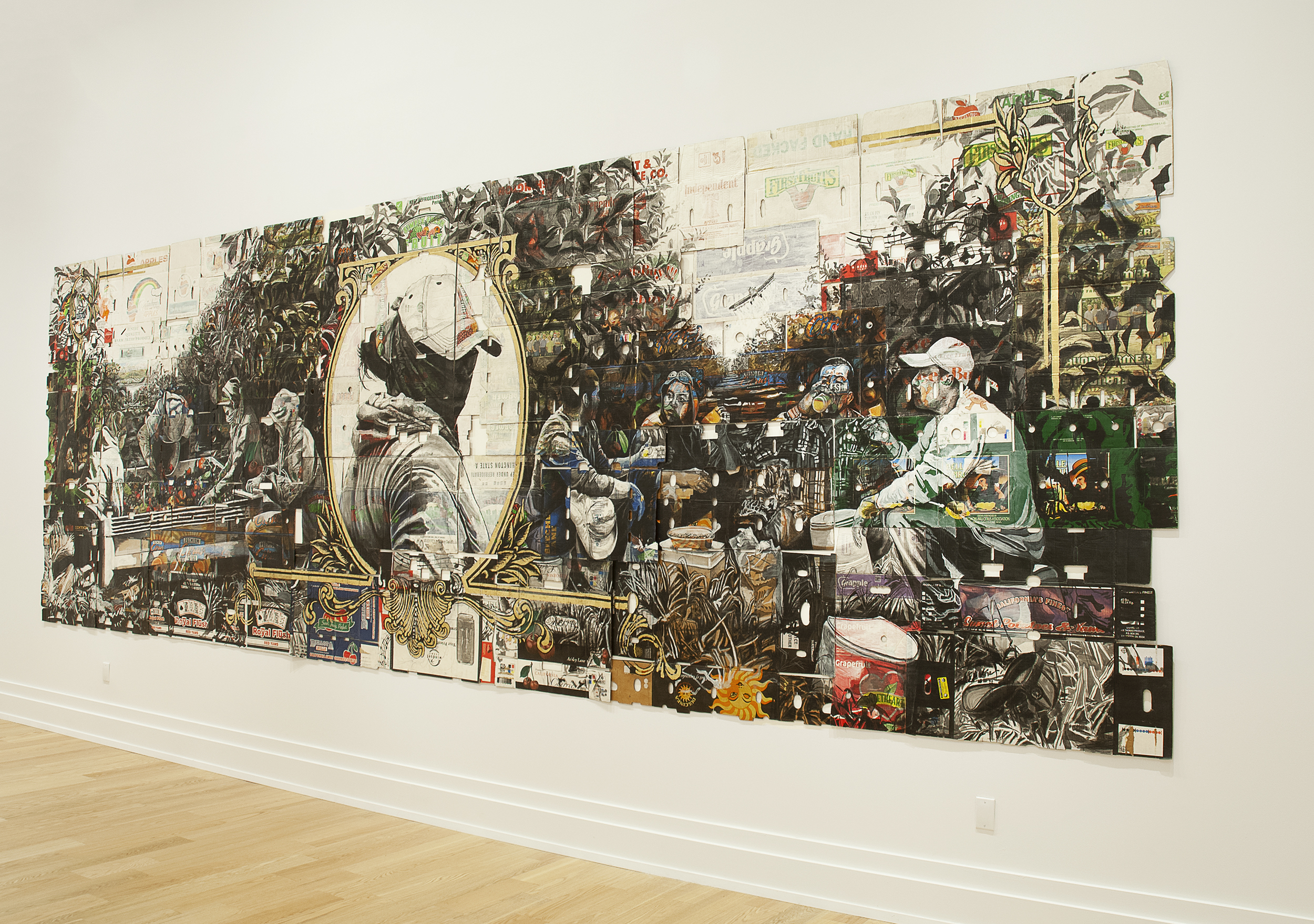 A large artwork takes up most of a wall, mimicking the look of a dollar bill but with imagery calling to agricultural labor in black ink and subdued colors, made on cardboard boxes