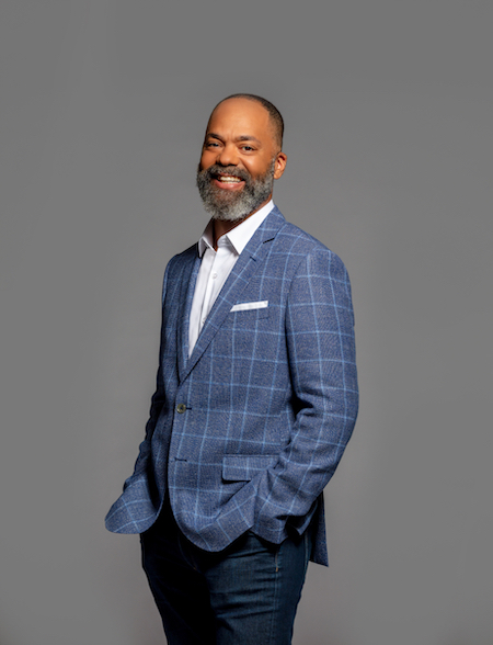 A middle aged man with medium dark skin tone and a grey beard smiling, wearing a blue plaid sport coat and jeans
