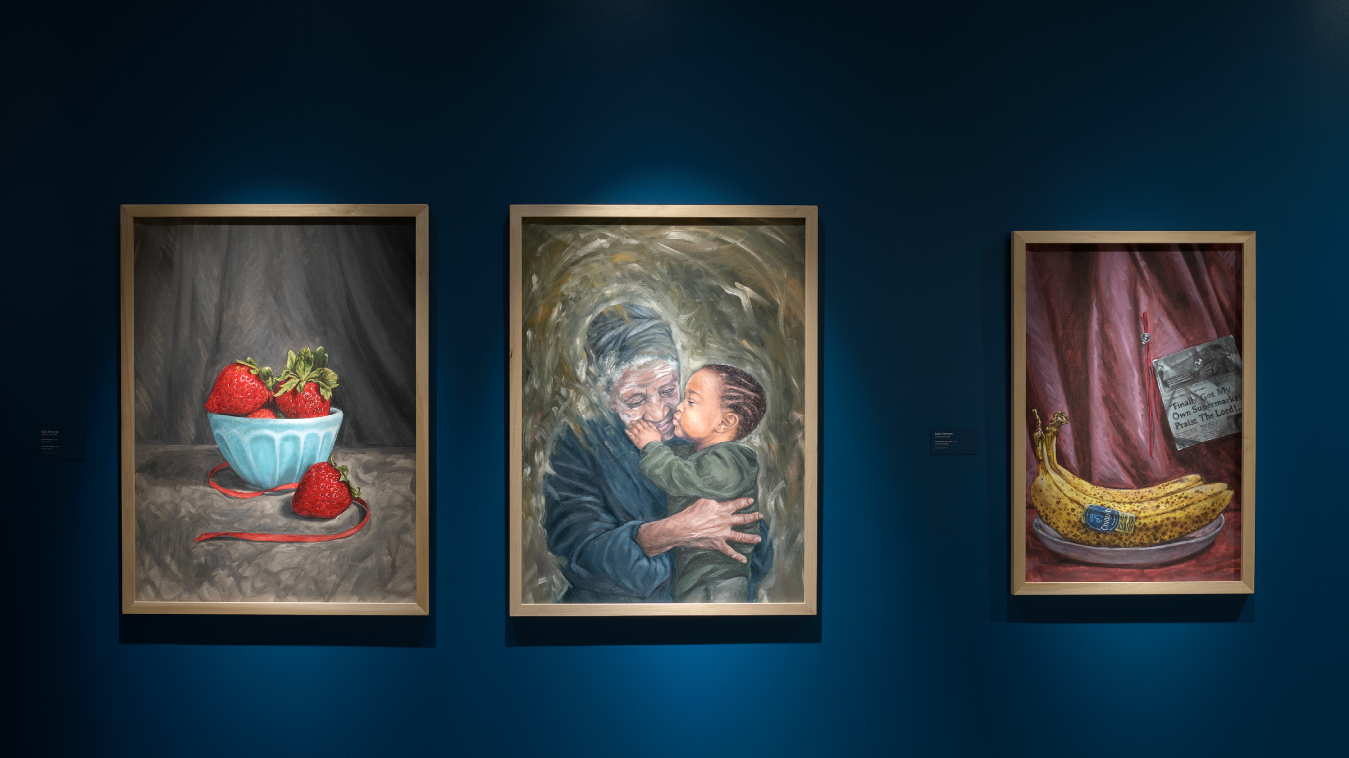 Installation views of three oil paintings in light wooden frames on a blue wall, the left being a still life of strawberries in a blue bowl, the middle a portrait of an older woman with dark skin tone embracing a young toddler with medium to dark skin tone wearing a green hoodie, and the right being a still life of two bananas with a newspaper clipping behind them