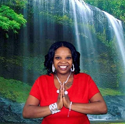 Headshot of a Black woman with her hands pressed together in front of her chest. There is a large photo of a waterfall behind her.