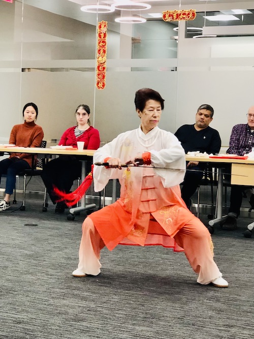 A woman in orange and white traditional Tai Chi robes practicing with a thin sword