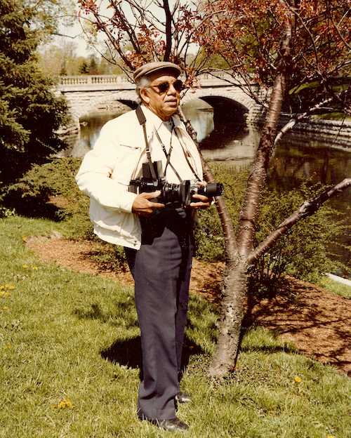 An older man of medium dark skin tone wearing a camera around his neck and white jacket in a green park