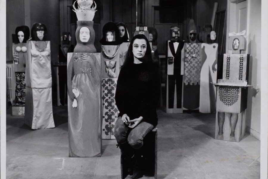 A black and white photograph of a Latinx woman wearing a black shirt and jeans and seated on a wooden box. In the background of several sculptures of people made from wood.