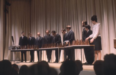 Nine men and one woman standing behind a long table with metronomes on it on the Auditorium stage