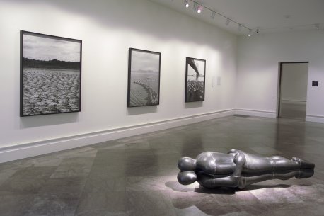 Installation view of three black and white photographs on the wall and a sculpture of two figures lying down on the floor, embracing