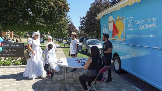 Two women in long white dresses stand with a child and another woman in front of an artmaking table in front of a large blue truck