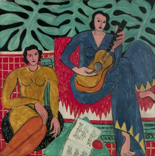 Two women sitting in front of lush green leaves. The woman in the yellow dress sits on a cushion on the floor while the woman in a blue dress sits in a red chair, holding a guitar.
