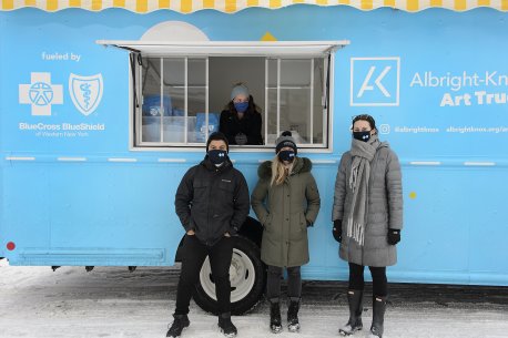 Three adults standing outside a big blue truck and one adult visible through the window of the truck