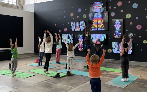 Kids doing yoga in front of an artwork of an artwork made up of several TV screens hanging on a black wall that show a colorful being