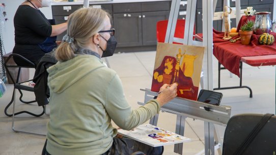 A woman working on a painting of a still life on a canvas on an easel in front of her