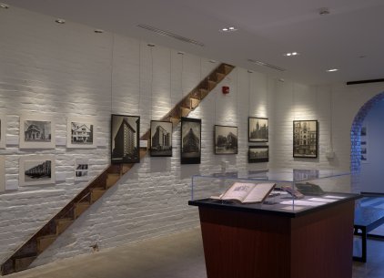 Black and white photographs of buildings hanging on a white stone wall, and a pedestal with books inside a case standing in the middle of the room