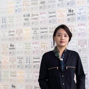 Mizin Shin, a Korean woman, stands in jacket in front of wall of her prints in multiple colors of &quot;Use Your Voice #StopAsianHate&quot;