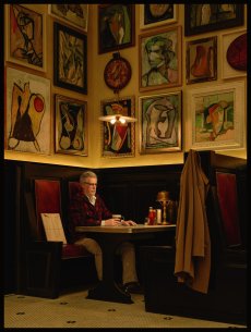 A man sitting at a booth in a restaurant with many paintings on the wall behind him