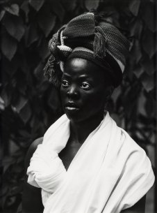 A Black woman wearing a dark colored headwrap and a white wrap around her shoulders standing in front of a wall of leaves
