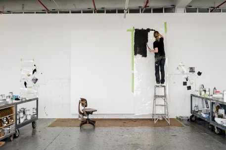 A woman on a ladder applying black paint to a large white paper taped to the wall