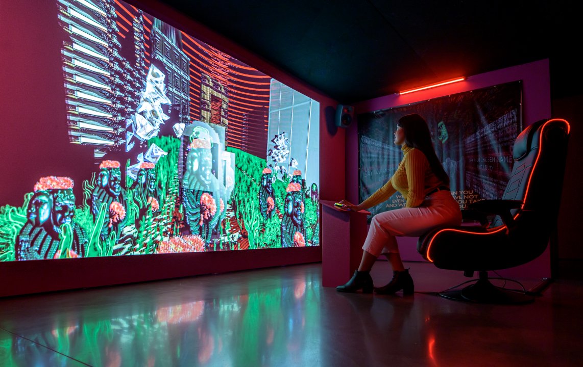 Installation with online game for one player, gaming chair, two pink fluorescent lights, and two hanging vinyls.