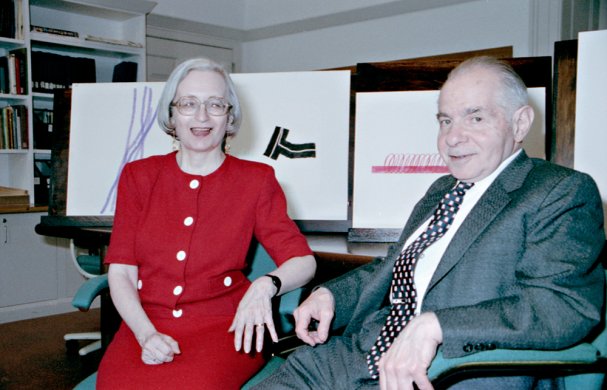 An older woman and man sitting next to each other in chairs with artworks behind them