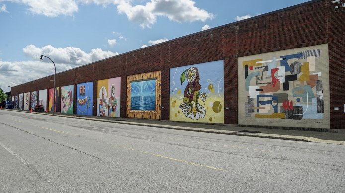 A receding line of varied murals on a long brick wall with blue, cloudy sky above.