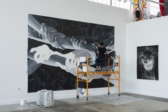 A woman with brown hair working on a large black and white painting of hands hanging on a large white wall