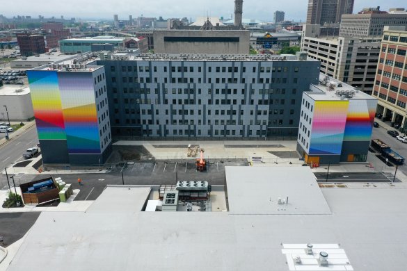 An elevated photograph of Josef Kristofoletti&#039;s Golden Hour, a mural that covers four faces of the building at 201 Ellicott Street in bands of bright color. In the background is the cityscape of Buffalo.