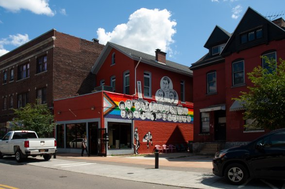 Mural of the faces of LGBTQ+ icons against the Progress Pride flag on a red brick building with city street and blue sky in background.