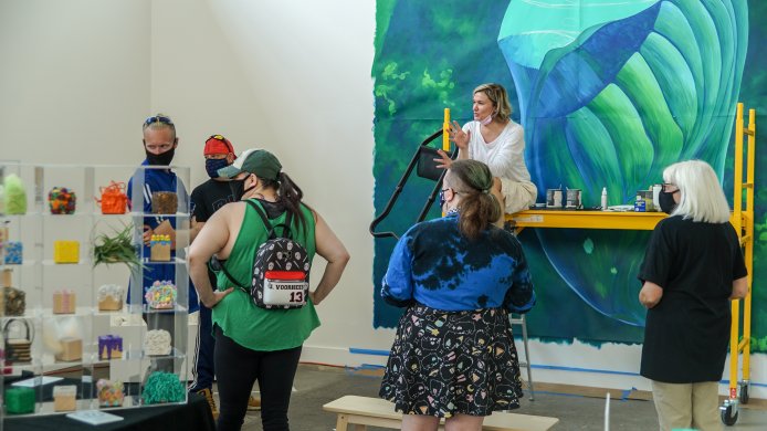 A woman sitting on yellow scaffolding in front of a large blue and green painting in progress talks to a group of adults standing on the floor in front of her
