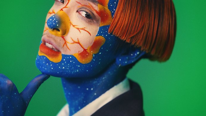 A person with a celestial designed facepaint and blue paint around their neck and hand with a red wig
