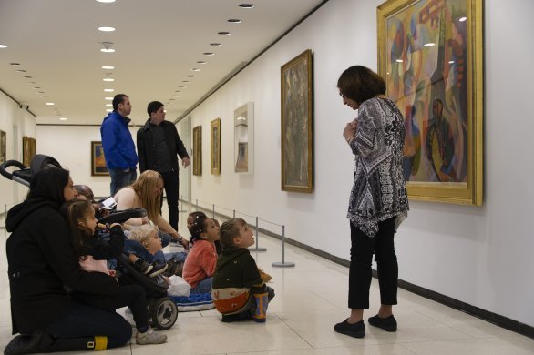 Families with young children sit on the floor in front of a large painting on the wall; a docent stands in front of the painting, talking