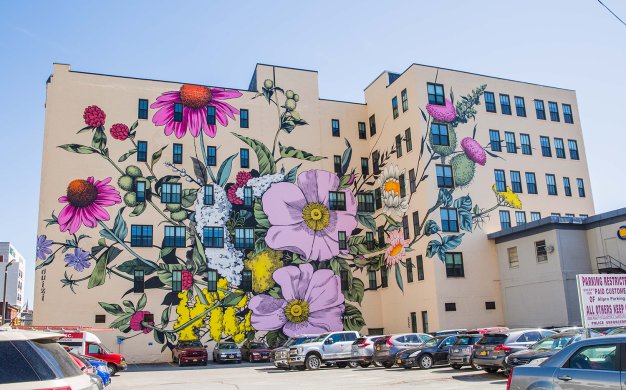 Giant flowers in several shades of pink, as well as purple, yellow, and white and their green stems and leaves cover much of the façade of a six-story building. A parking lot with cars in front of the building is visible in the foreground of the image.