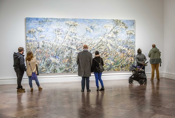 Six adults and one kid in a stroller in front of a large painting with a blue sky and wildflowers