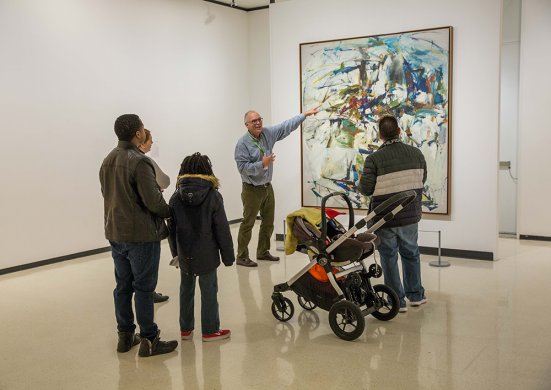 Two adults, a kid, and a baby in a stroller standing in front of a docent, who is gesturing toward an abstract painting featuring blue and green strokes