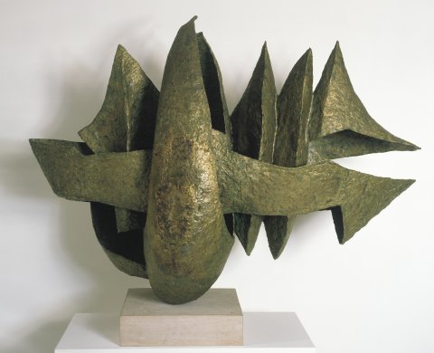 Bronze-colored, jagged vertical forms grouped together appear to balance on top of a small, square, stone base. The majority of the sculpture extends to the right, narrowing to a sharp point resembling the tip of a spear. Five jagged tips point upward, and three on the right side point down. Each section is angular, casting dark shadows throughout the sculpture, and the bumpy surface throughout adds an aged, or patina-like appearance.