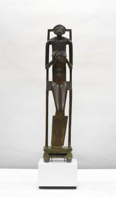 A life-sized bronze abstract female nude holds her hands placed in front of her chest, as if cradling an invisible sphere. She appears to be perched on a tall chair, which supports and frames the figure from behind. The facial features on her flat, round head are angular. Her long, skinny body terminates in long legs bent at the knee. The figure’s feet rest on a narrow rectangular shaped stool at the sculpture’s base.