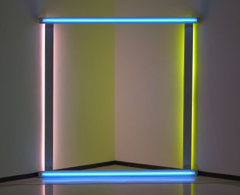 A square of fluorescent tube lights mounted in white aluminum fixtures sits on the floor in front of a corner of the gallery space. The lights that make up the top and the bottom of the frame shine bright blue light outward. The lights that make up the right and left of the frame cast yellow and pink light into the corner behind the sculpture.