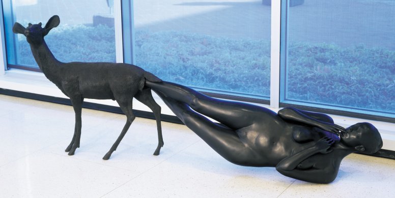 A life-sized nude female figure emerges head-first from the rear of a standing deer. Only her feet remain inside the animal, and the rest of her body is angled toward the floor, with her left shoulder making contact. She cranes her neck slightly to gaze back toward the deer, who has large ears and small antler nubs. The bronze surface of the sculpture is smooth and has a dark uniform patina, making it appear black.