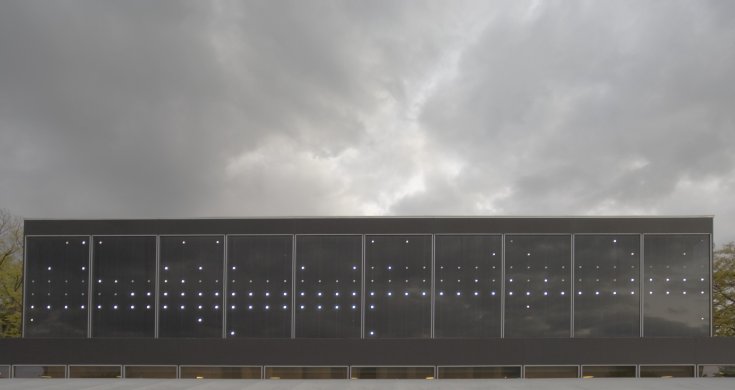 Small white dots of varying brightness speckle a wall of ten panels of dark-colored glass as seen from the outside. These dots seem to fit into a matrix of horizontal and vertical rows but without a discernable pattern. This wall of windows only fills the bottom half of the image, and a sky filled with ominously dark clouds is visible above.