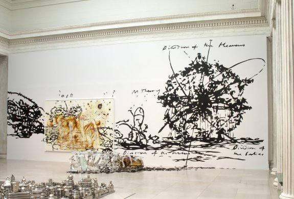 Black, vine-like tendrils cover much of a large white wall. These are interrupted to the left of center by a large painting featuring more of the tendrils overlaid with abstract washes of copper and taupe paint. On the right side, the tendrils coalesce in a large sphere encircled by the phrases “division of the heavens,” “division of the waters,” “division of the bodies,” and “M theory” in cursive. On the floor in front of the wall rests a long, tube of perforated aluminum surrounded by Tarot cards.