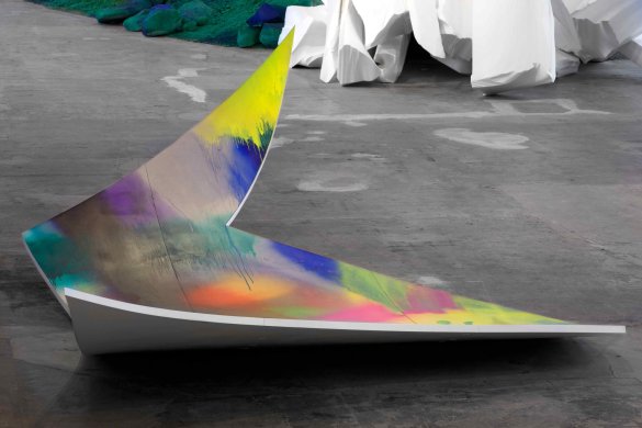 This abstract sculpture appears overall like an oversized piece of paper with a V-shaped divot cut out of the right side and curling edges. It balances on the floor near its center. The underside of the plastic form is white, but bright patches of spray-painted yellow, blue, green, purple, orange, black, gray, and pink cover the top, overlapping and dripping onto one another.