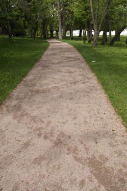 One of five images. This vertically oriented photograph shows a landscape with patches of lush green grass separated by a graveled walking path stretching into the distance and toward a cluster of trees. A slightly darker brown wave pattern zigs and zags across the width of the pathway, running along its full length.