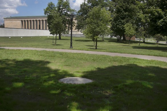 A flat circle of stone perforates the grass in the center of this image’s lower foreground. A portion of the museum’s neoclassical building is visible at top left, and trees largely fill the center and right-hand portion of the background.