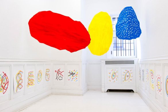 Pieces of white paper with colorful paintings on them fastened to a white wall. Large red, yellow, and blue shapes hang from the ceiling.