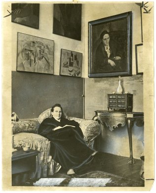 A black and white photograph of a woman sitting on a cough with paintings hanging on the wall behind her