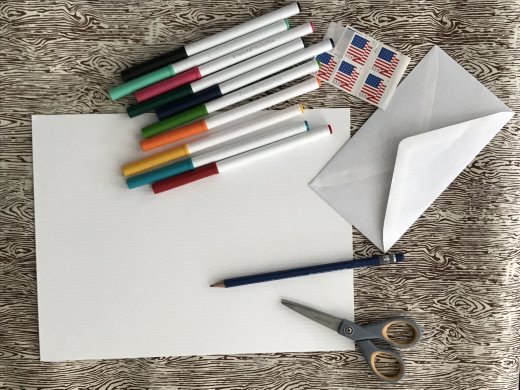 A bird's eye view of artmaking supplies, including white paper, markers, a pencil, scissors, an envelope, and stamps