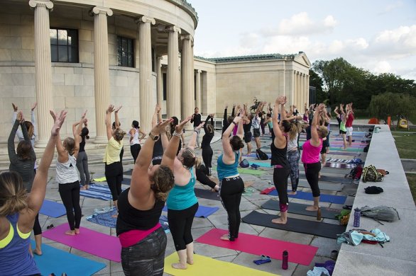 Adults standing on yoga mats with their arms up in the air, outside on the Elmwood Avenue Portico